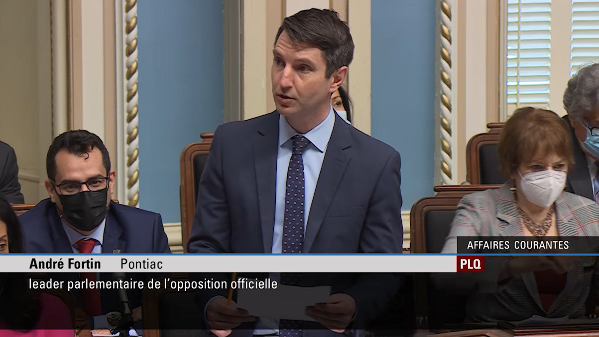 Le député du Parti libéral du Québec, André Fortin, s’exprime debout à l’Assemblée nationale du Québec, tenant des documents lors d’une période de débats parlementaires. D’autres élus sont visibles assis derrière lui, dans un cadre institutionnel formel.