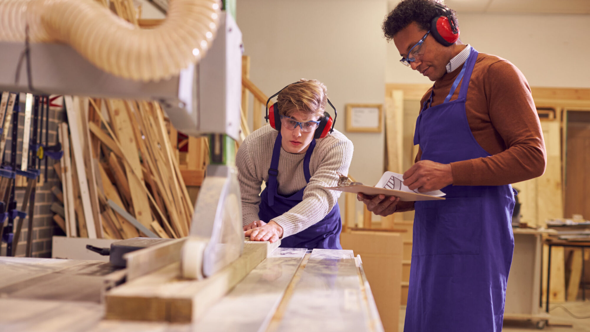 Tutor With Male Carpentry Student In Workshop Studying For Apprenticeship At College Using Bench Saw