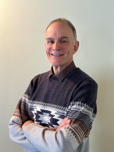 Photo de face Jocelyn Carle sur mur beige avec chemisier et chandail bruns