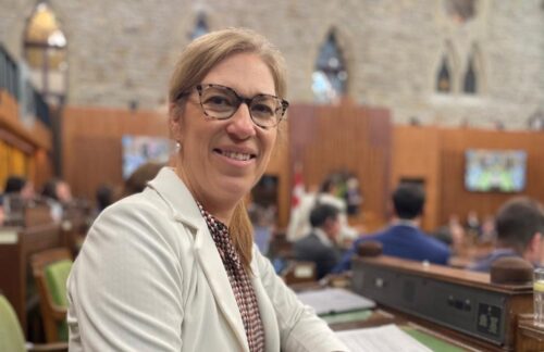 Une femme avec des lunettes et un veston blanc, assise, regarde vers l'objectif. En arrière-plan, on voit des bancs et des gens lors de ce qui ressemble à une session parlementaire.