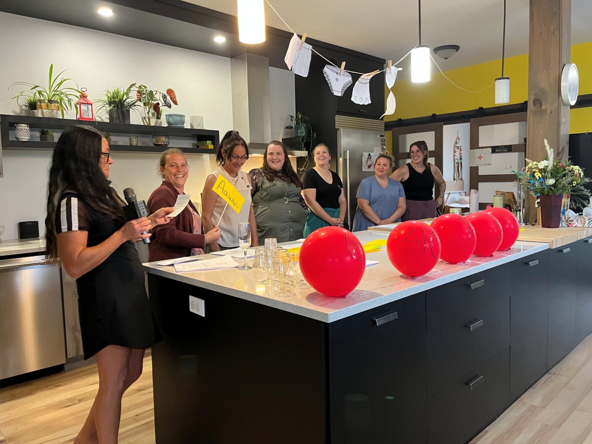 7 femmes sont debout derrière le comptoir noir et blanc d'une cuisine de style moderne. Elles rient toutes et l'une d'entre elles tient un micro et une feuille blanche. 5 ballons à l'hélium reposent sur le comptoir.