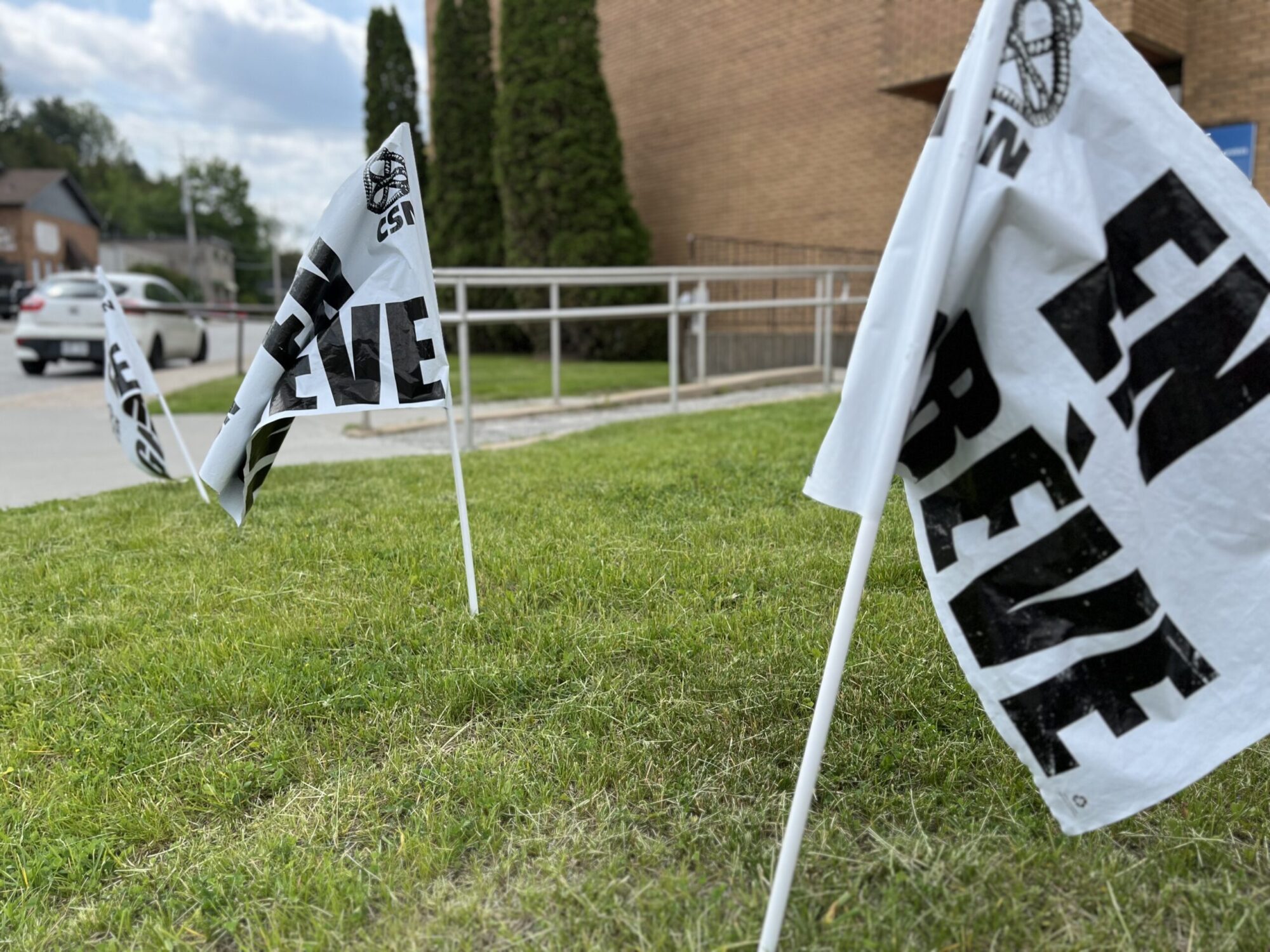 De petits drapeaux blancs avec le slogan " en grève " imprimé dessus son plantés dans de la pelouse en bordure de route. à l'arrière, on aperçoit un bâtiment.