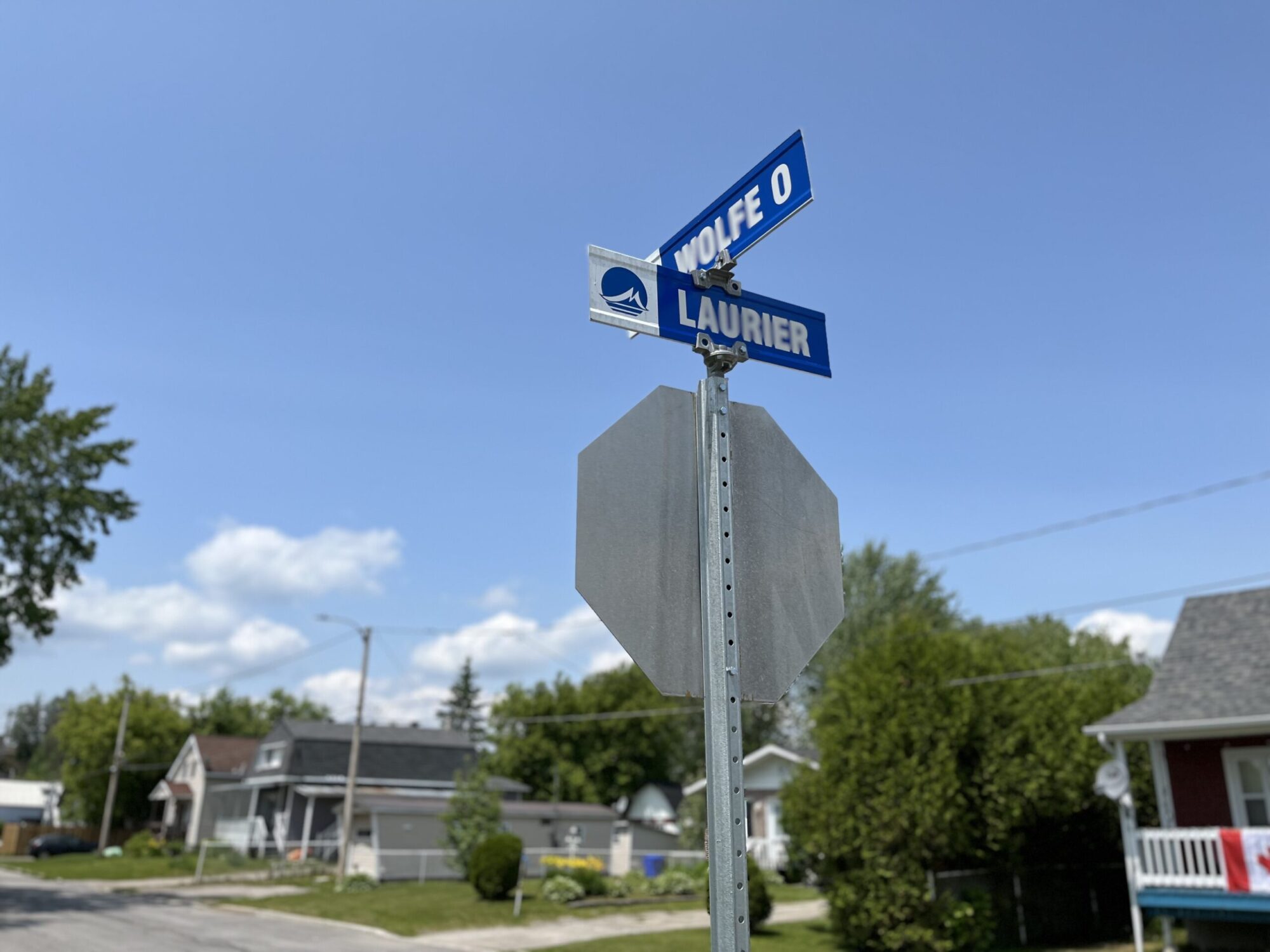 Un panneau arrêt vu à l'envers et des panneaux de rues municipales indiquant les rues Laurier et Wolfe. En arrière, une rue, des arbres et des maisons dans un quartier résidentiel sous un ciel bleu.