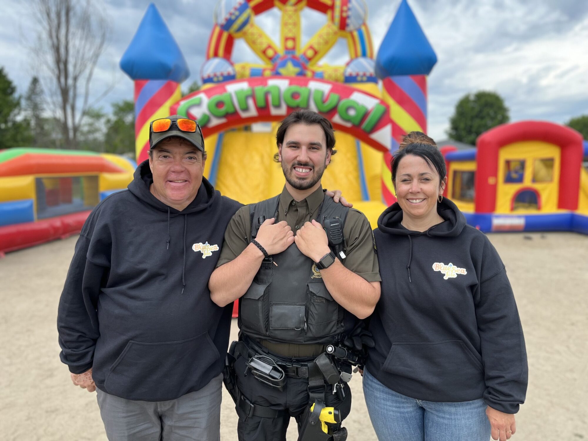 Un homme et une femme avec un haut noir de bénévole pour l'évènement Blue Sea en fête prennent la pose devant des jeux gonflables pour enfants, avec un policier de la Sûreté du Québec entre eux.