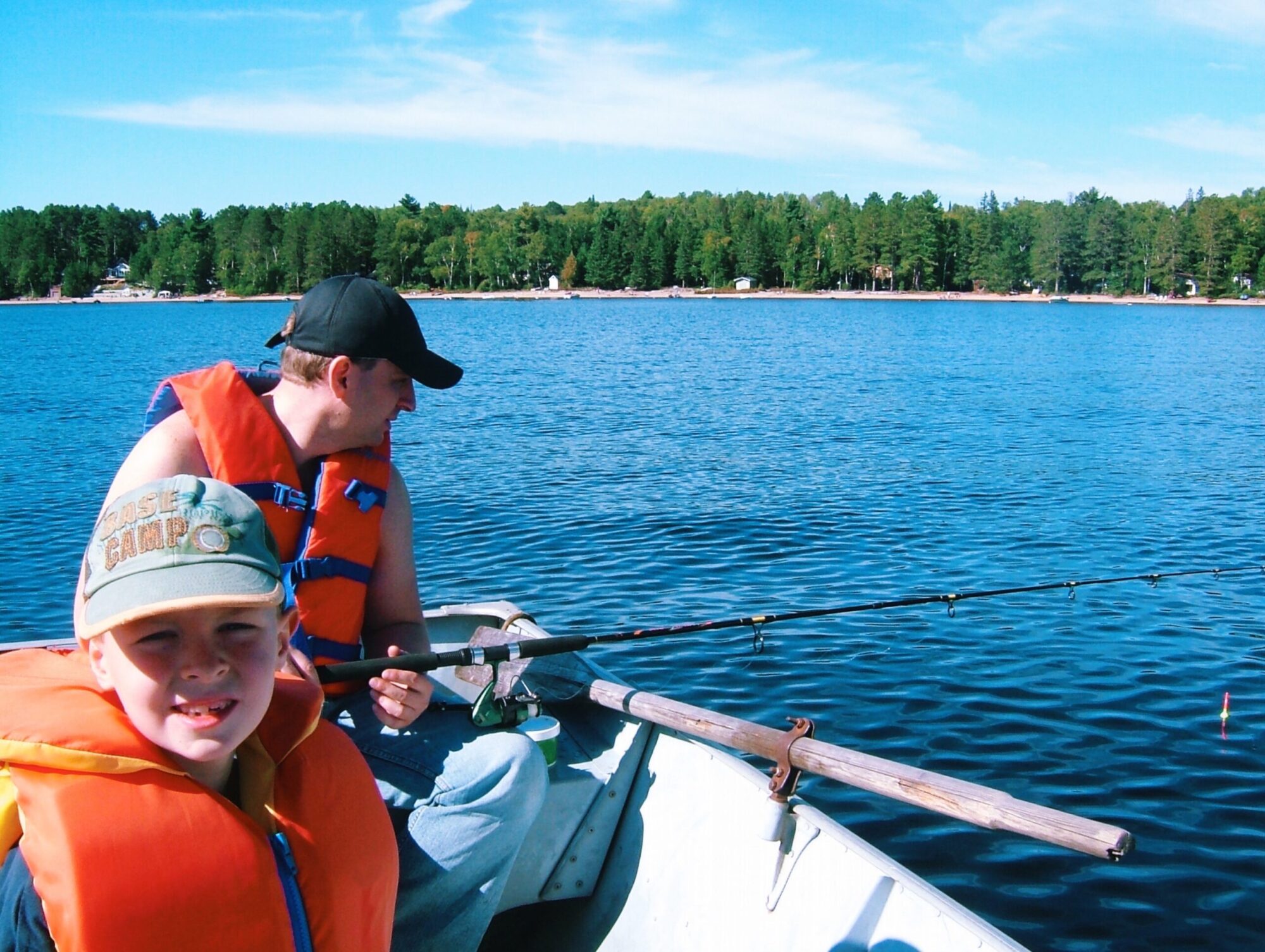 Un homme et un jeune garçon portant chacun une casquette et une veste de flottaison, pêchent depuis une embarcation sur un lac en été par beau temps.