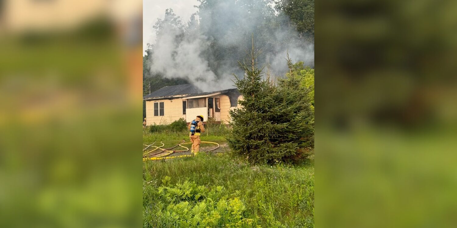Un pompier combat l'incendie d'une maison d'où s’échappent d’épais nuages de fumée en milieu boisé, sur un terrain herbeux.