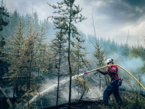 Un pompier forestier de la SOPFEU avec un casque blanc, vêtu d'une chemise rouge et d'un pantalon blanc, tient une lance à incendie dans ses mains alors qu'il étaient des flammes dans une forêt de résineux au Canada.