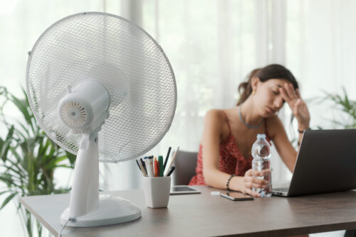 Une femme assise à un bureau devant son ordinateur tient une bouteille d'eau dans une main et son front dans l'autre, tandis qu'un ventilateur en fonction posé sur le bureau envoie de l'air dans sa direction.