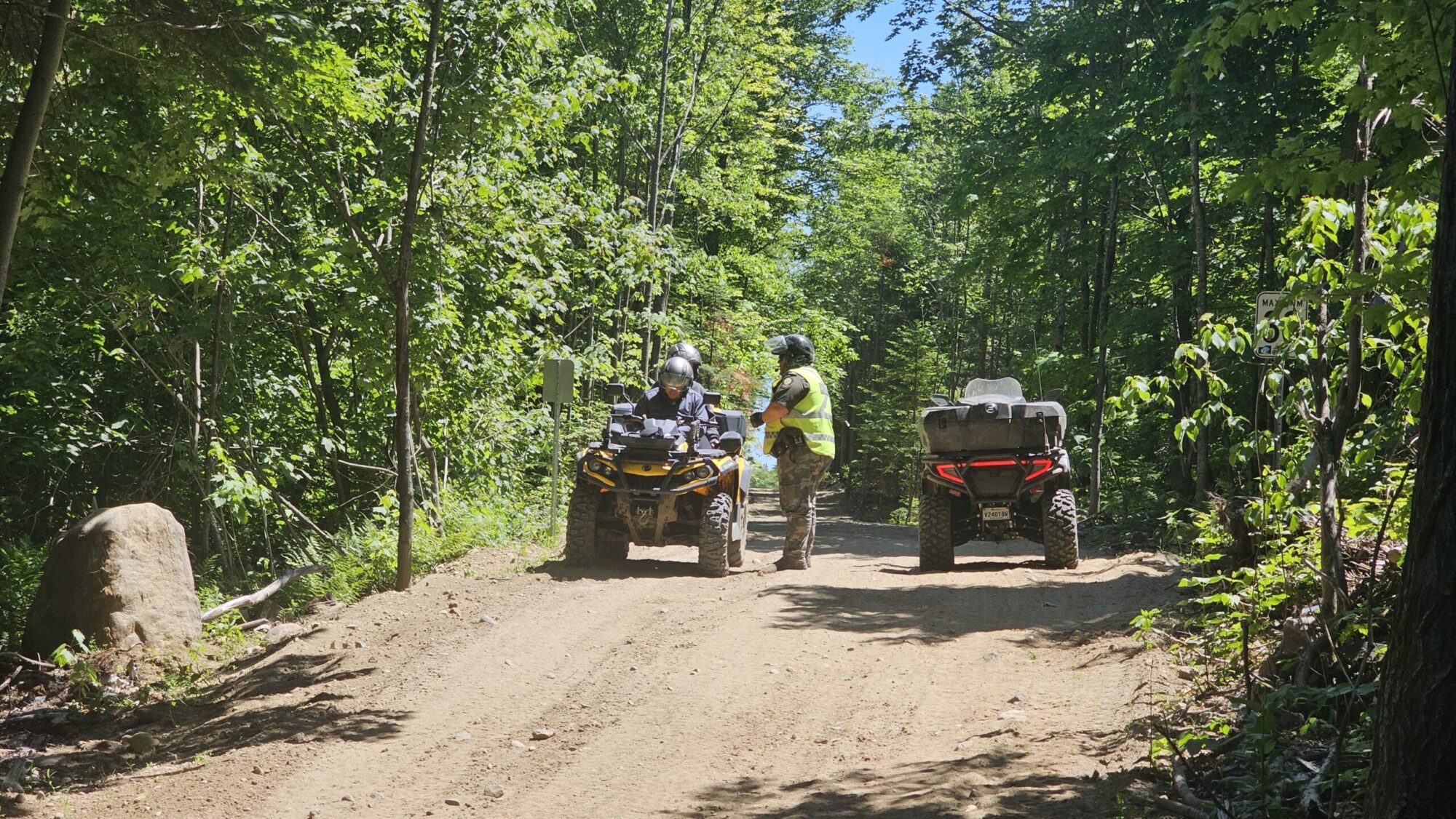 Un agent de la Sûreté du Québec avec un dossard jaune près de son véhicule hors route s'entretient avec des quadistes dans un sentier forestier.