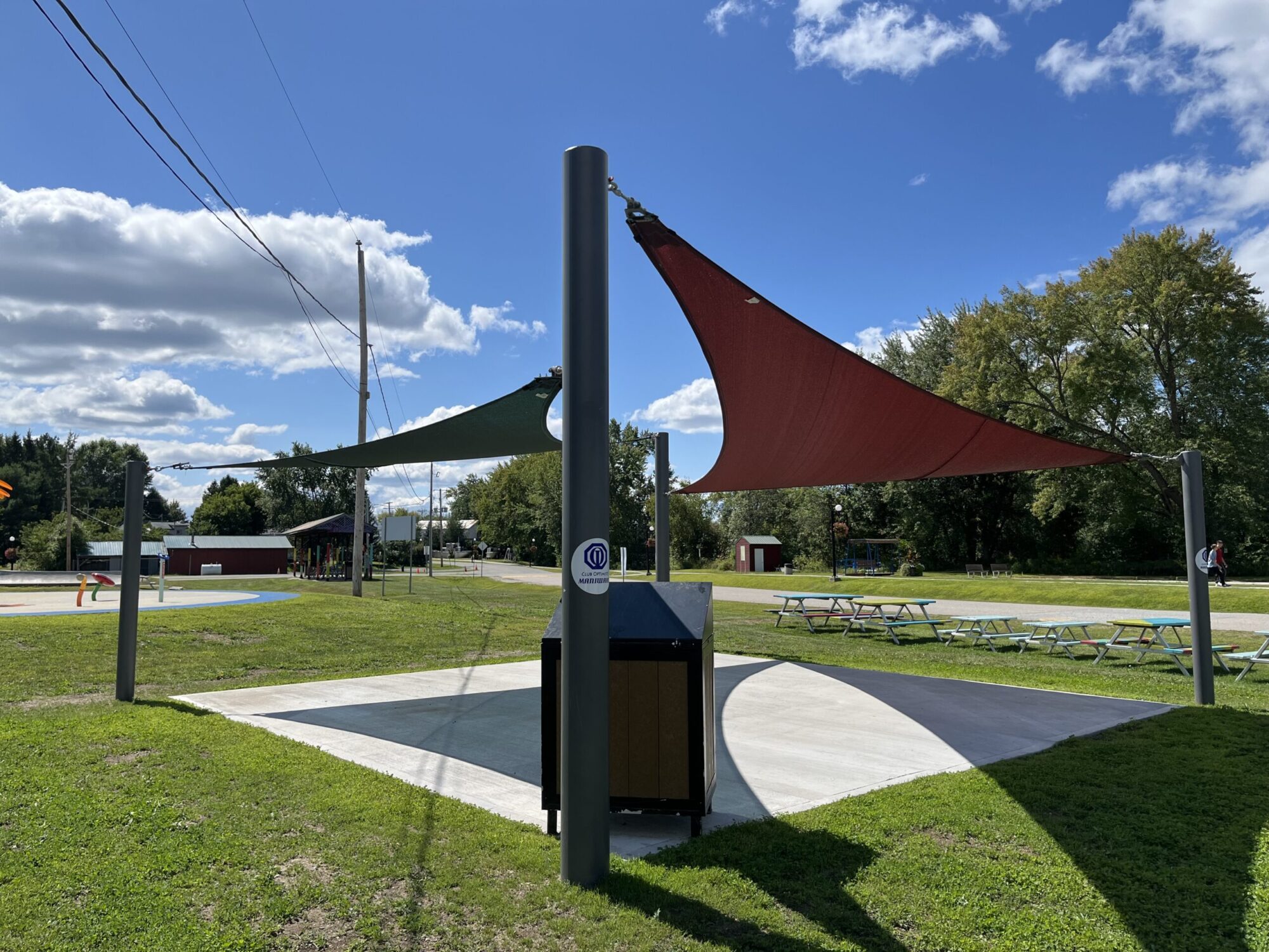 Un abri solaire fixé par des poteaux à une dalle de béton dans un parc municipal par une belle journée estivale.