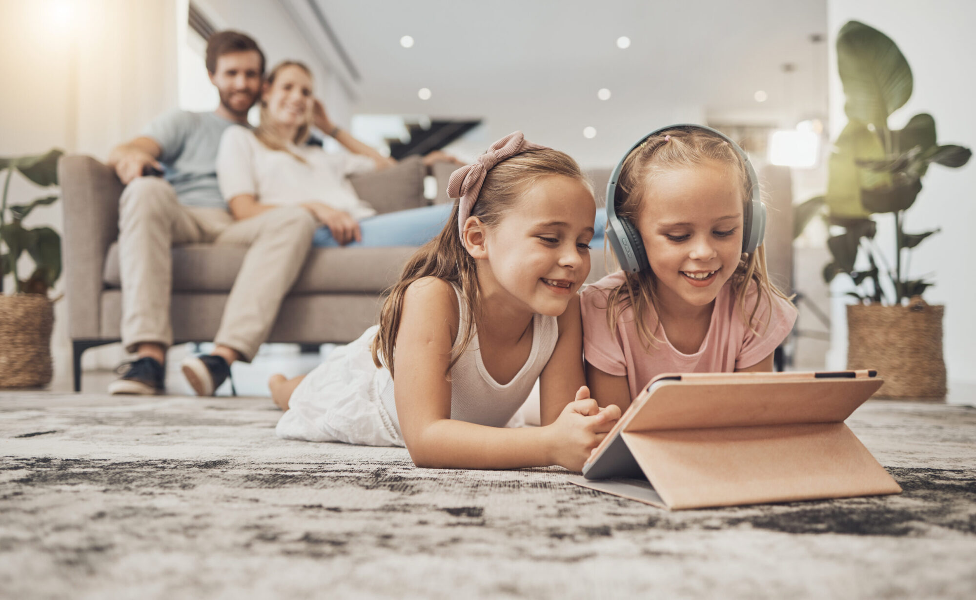 Deux petites filles regardent une tablette allongées dans un salon avec un couple adulte en arrière-plan assis sur un canapé.