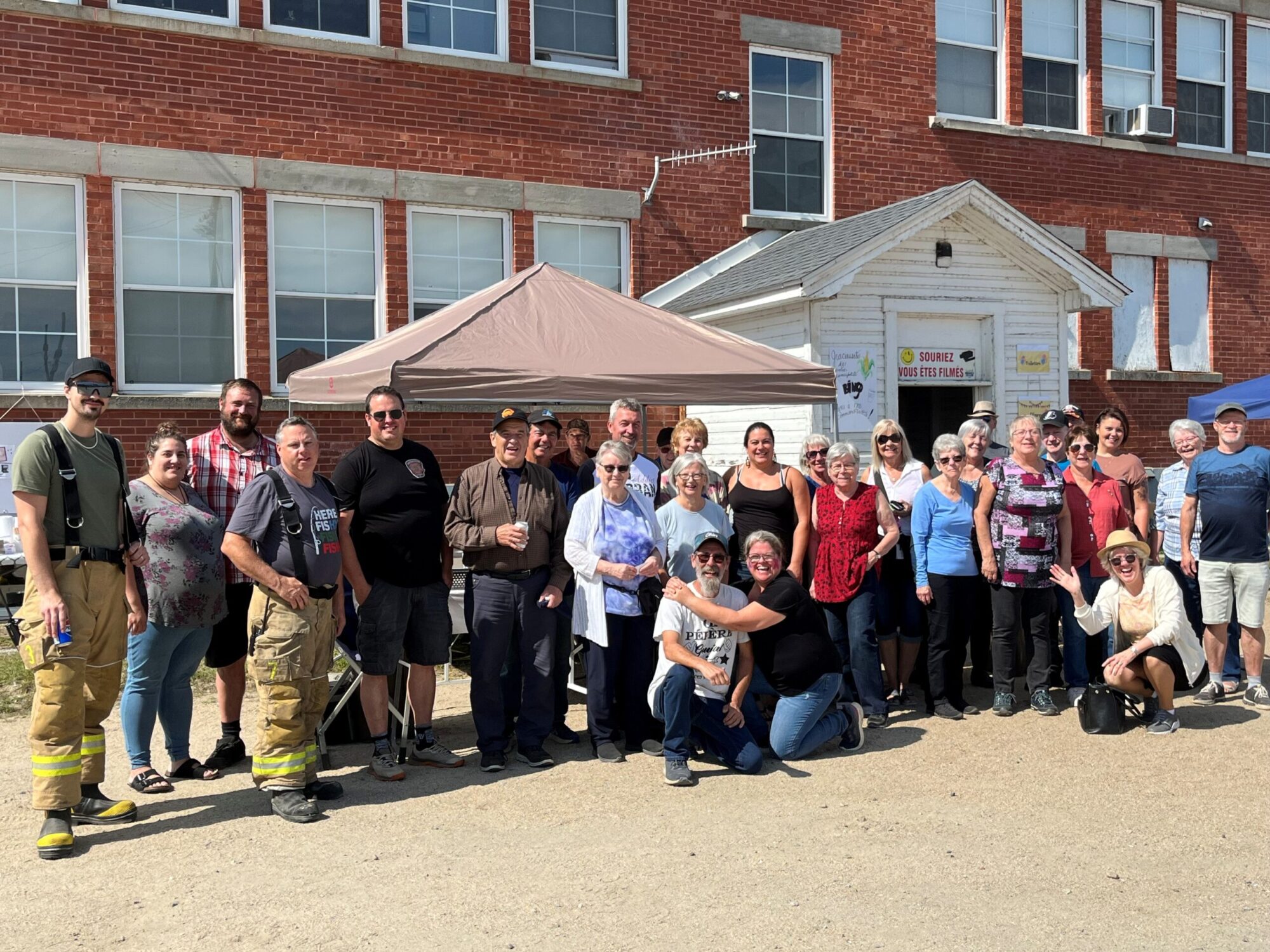Un groupe de personnes rassemblées devant un bâtiment en briques rouges, sous une tente installée pour l’occasion. L’ambiance est conviviale et festive.