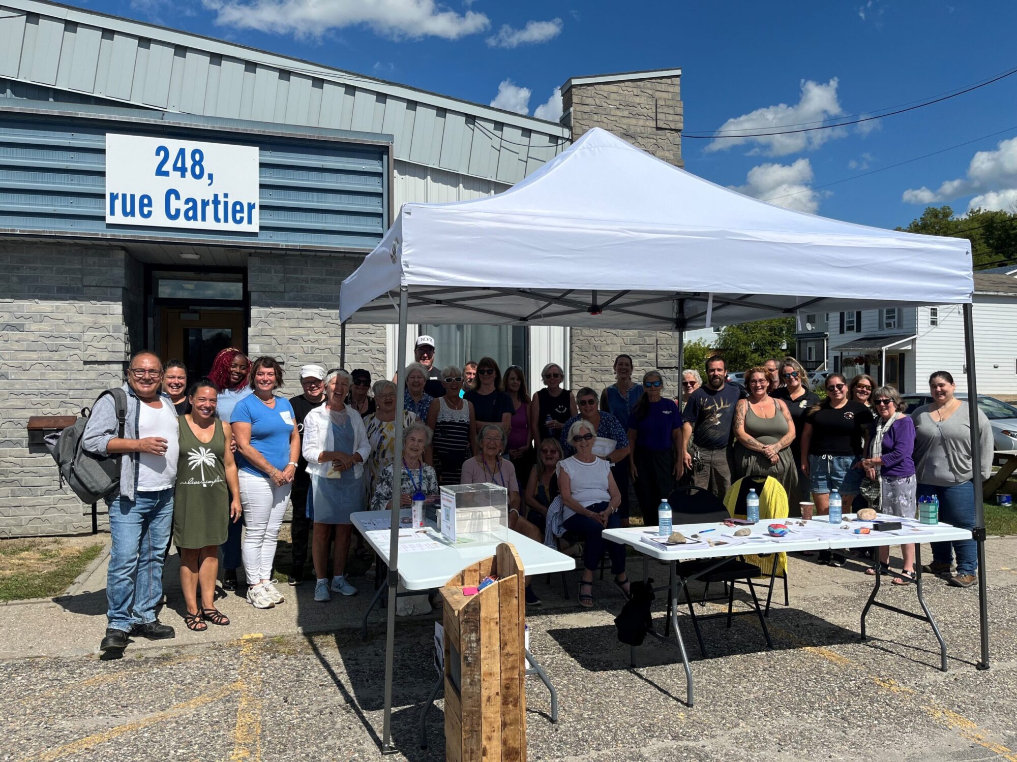 Un groupe de personnes réunies à l’extérieur d’un bâtiment situé au 248, rue Cartier, sous un chapiteau blanc et autour de tables installées pour l’occasion.