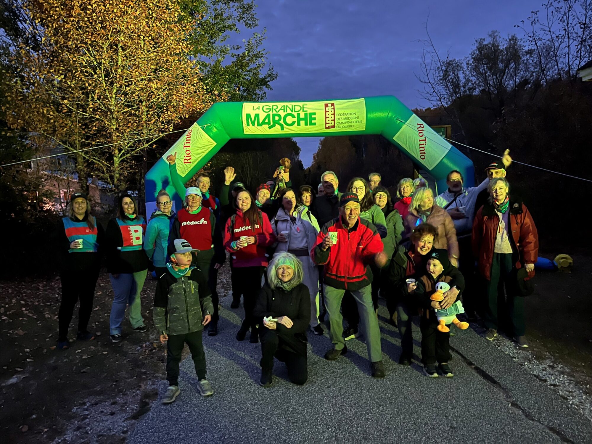 Un groupe de personnes de tous âges est rassemblé à l’extérieur devant une arche gonflable portant l’inscription « La Grande Marche ». Elles sourient et posent pour la photo dans une ambiance de fin de journée, avec un éclairage vert illuminant la scène.