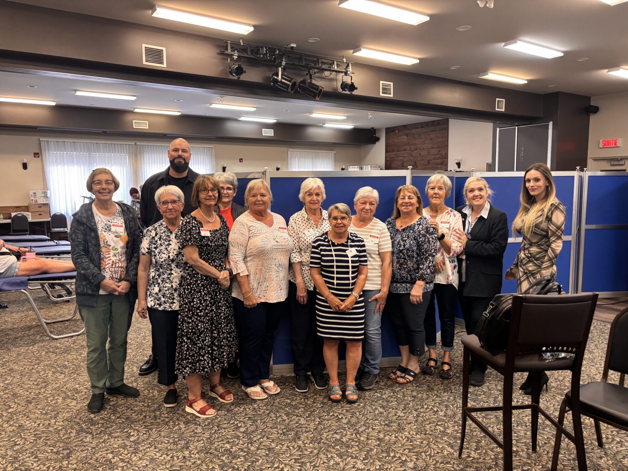 Un groupe de personnes posant à l’intérieur d’une grande salle. La majorité du groupe est composée de femmes, accompagnées d’un homme à l’arrière. On aperçoit des lits ou tables de repos en arrière-plan, ce qui suggère qu’il pourrait s’agir d’une collecte de sang.