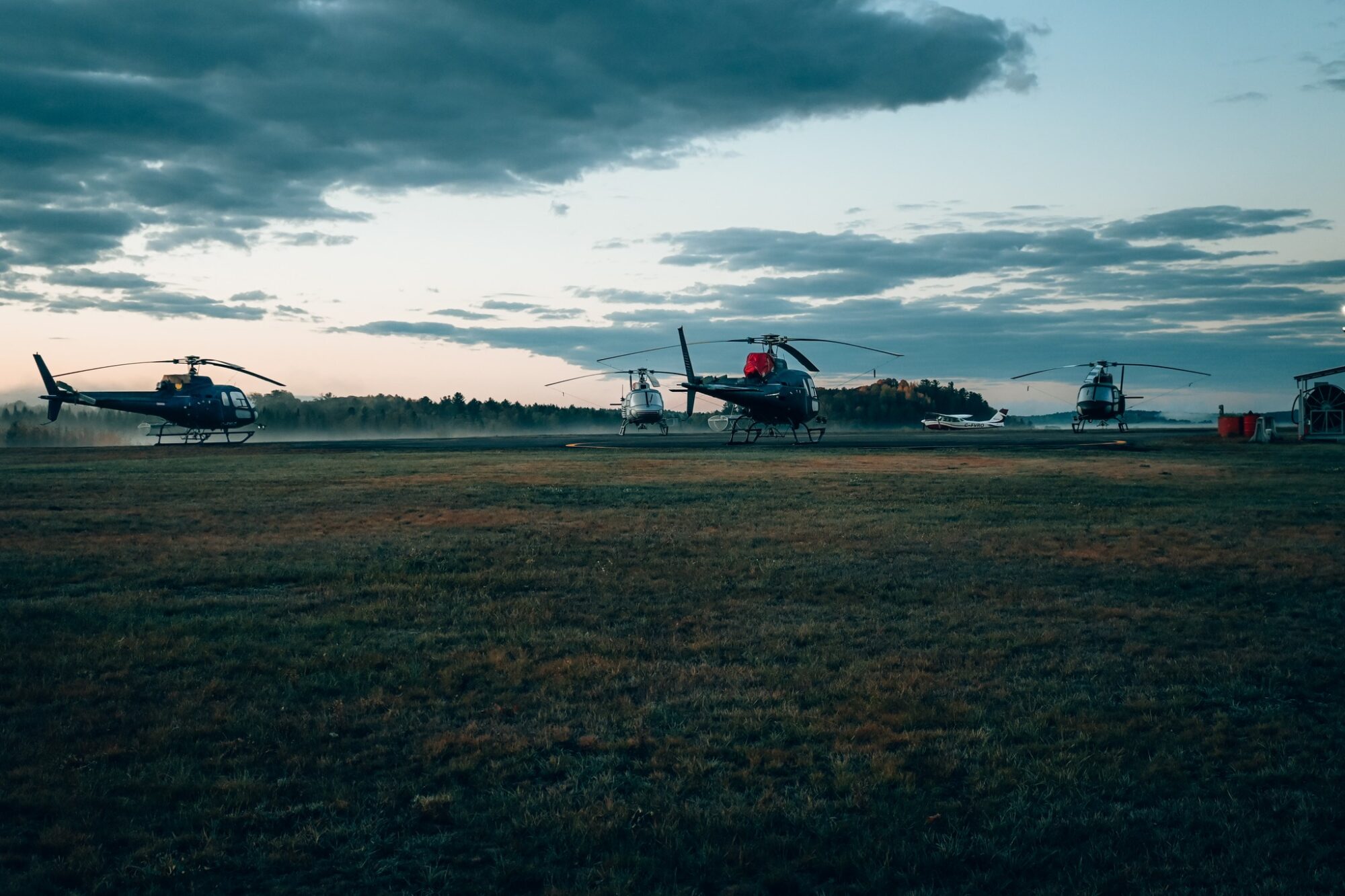 Quatre hélicoptères sont stationnés sur une aire herbeuse à ciel ouvert, au crépuscule. À l’arrière-plan, la lisière d’une forêt et un ciel nuageux complètent la scène.