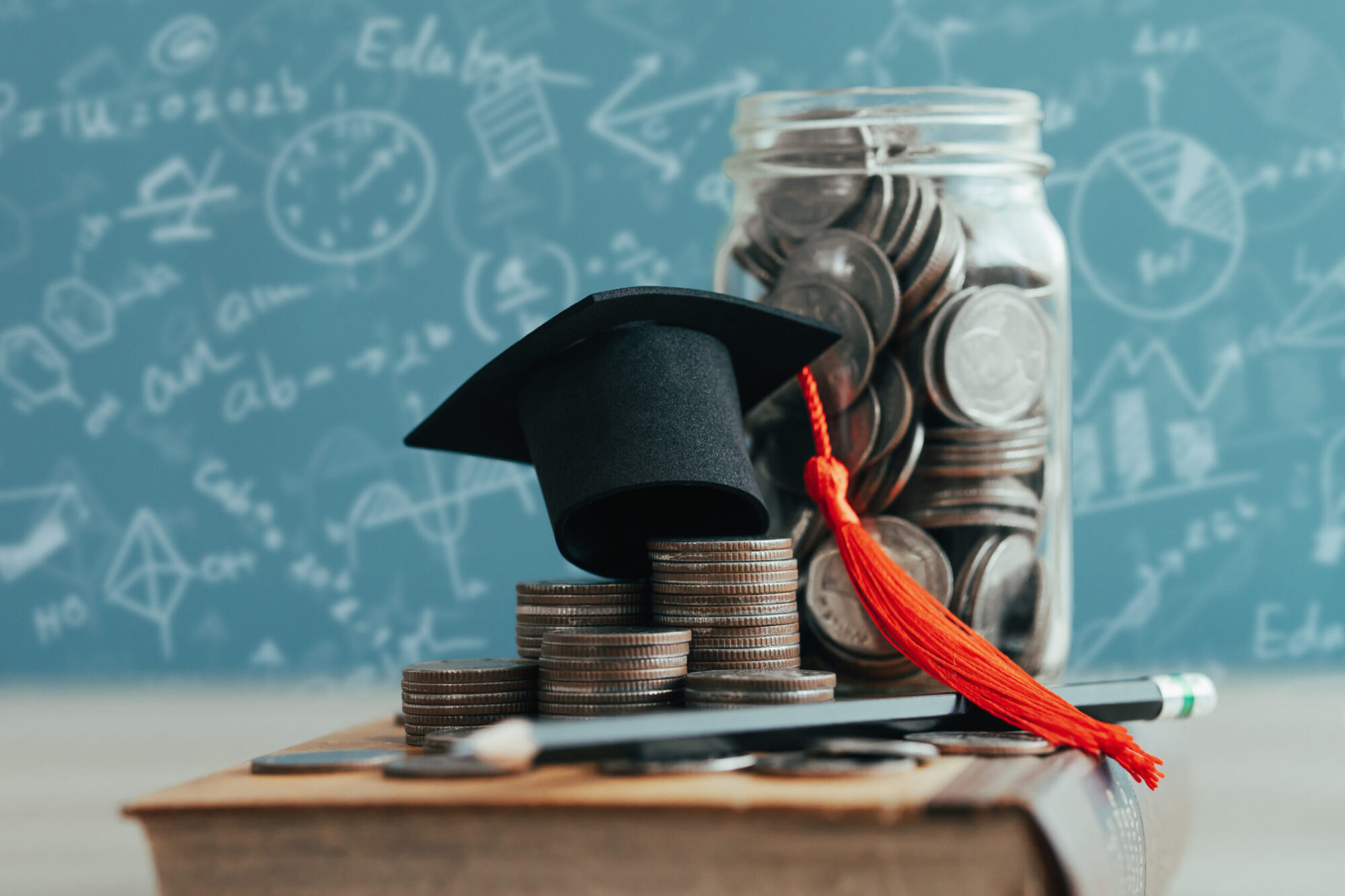 Une pile de pièces de monnaie surmontée d’un petit chapeau de graduation, placée à côté d’un pot rempli de pièces devant un tableau scolaire. Le tout est déposé sur un pupitre de classe d'école.