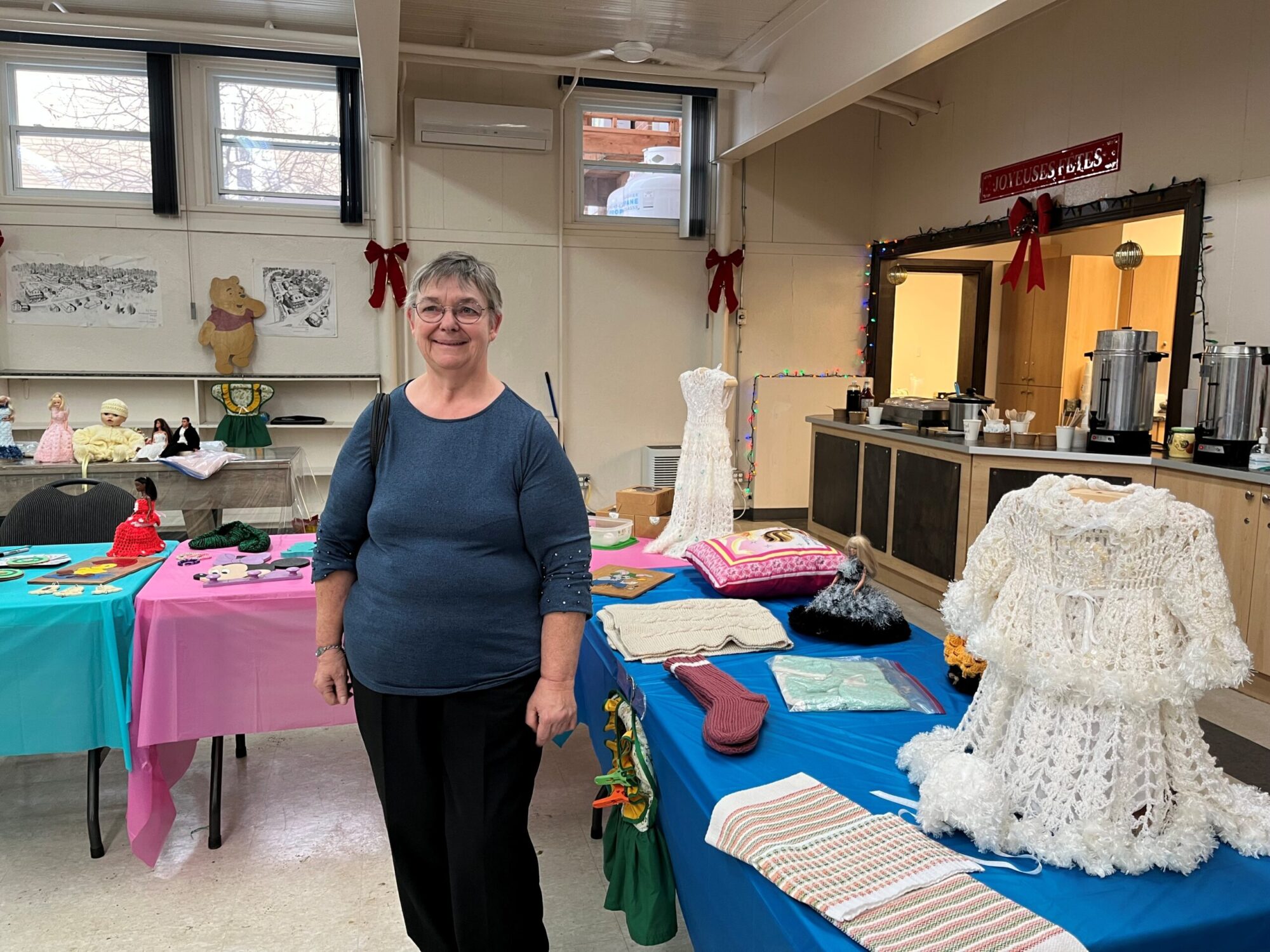 Une femme souriante se tient devant une table où sont exposés divers articles faits à la main, dont des vêtements au crochet, des poupées et des pièces artisanales colorées. L’ensemble est présenté dans une salle décorée pour le temps des Fêtes