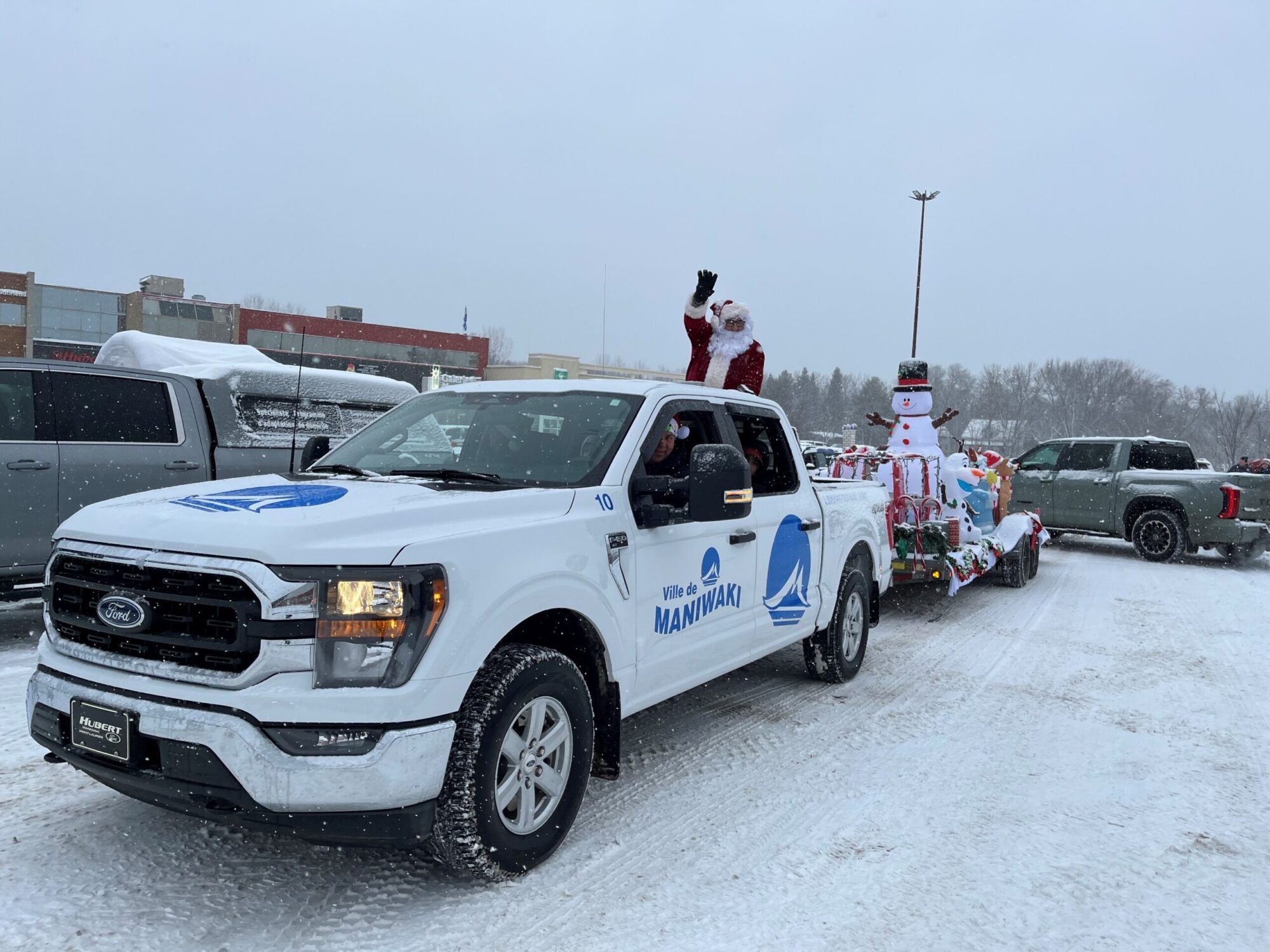 Un camion de la Ville de Maniwaki tire un char allégorique décoré pour Noël, avec un Père Noël debout à l’arrière qui salue la foule. La scène se déroule sous une neige légère, créant une ambiance hivernale typique d’un défilé festif.