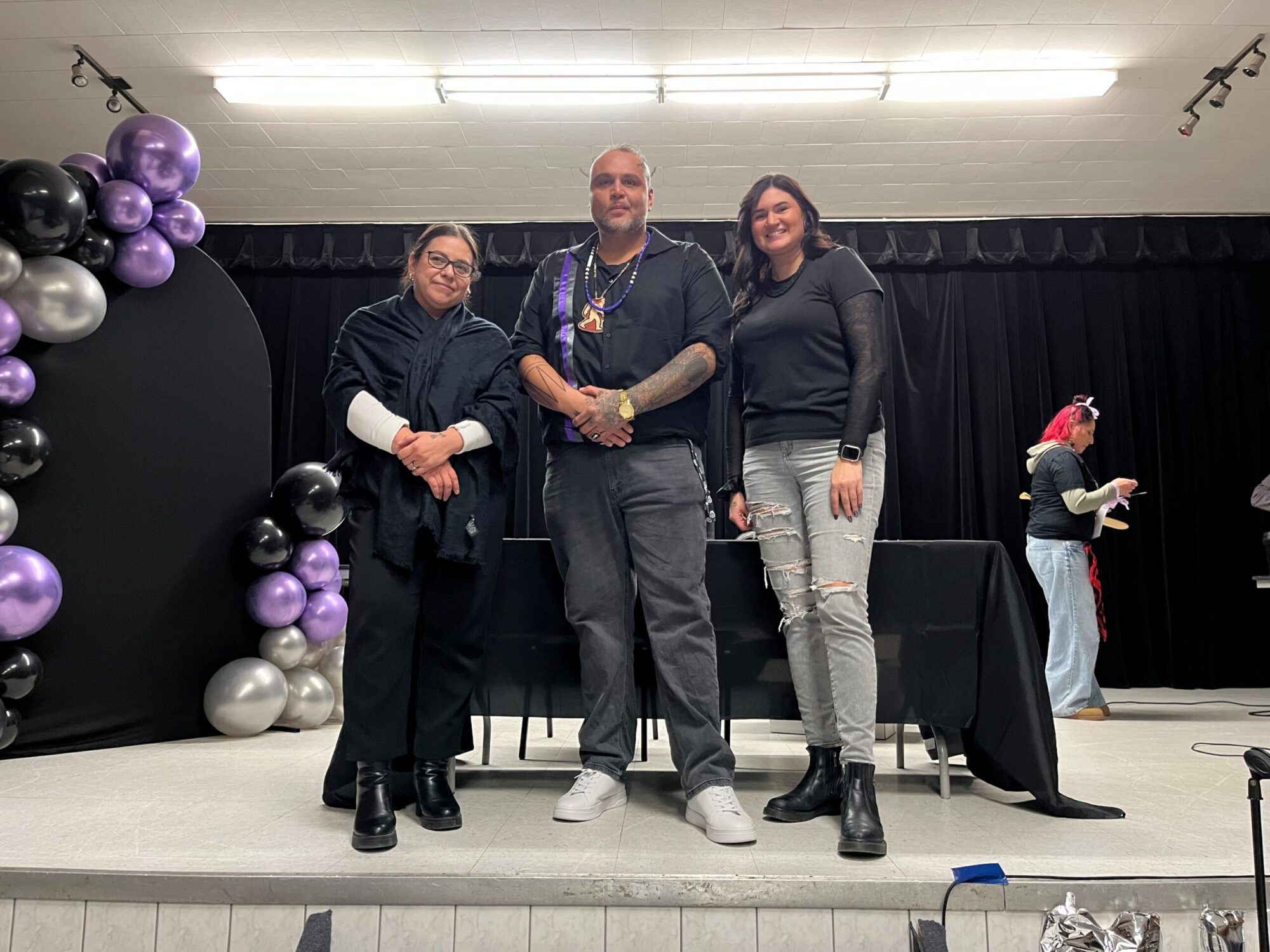 Trois personnes posent ensemble sur une scène décorée de ballons noirs, mauves et argentés, créant un décor festif et soigné.