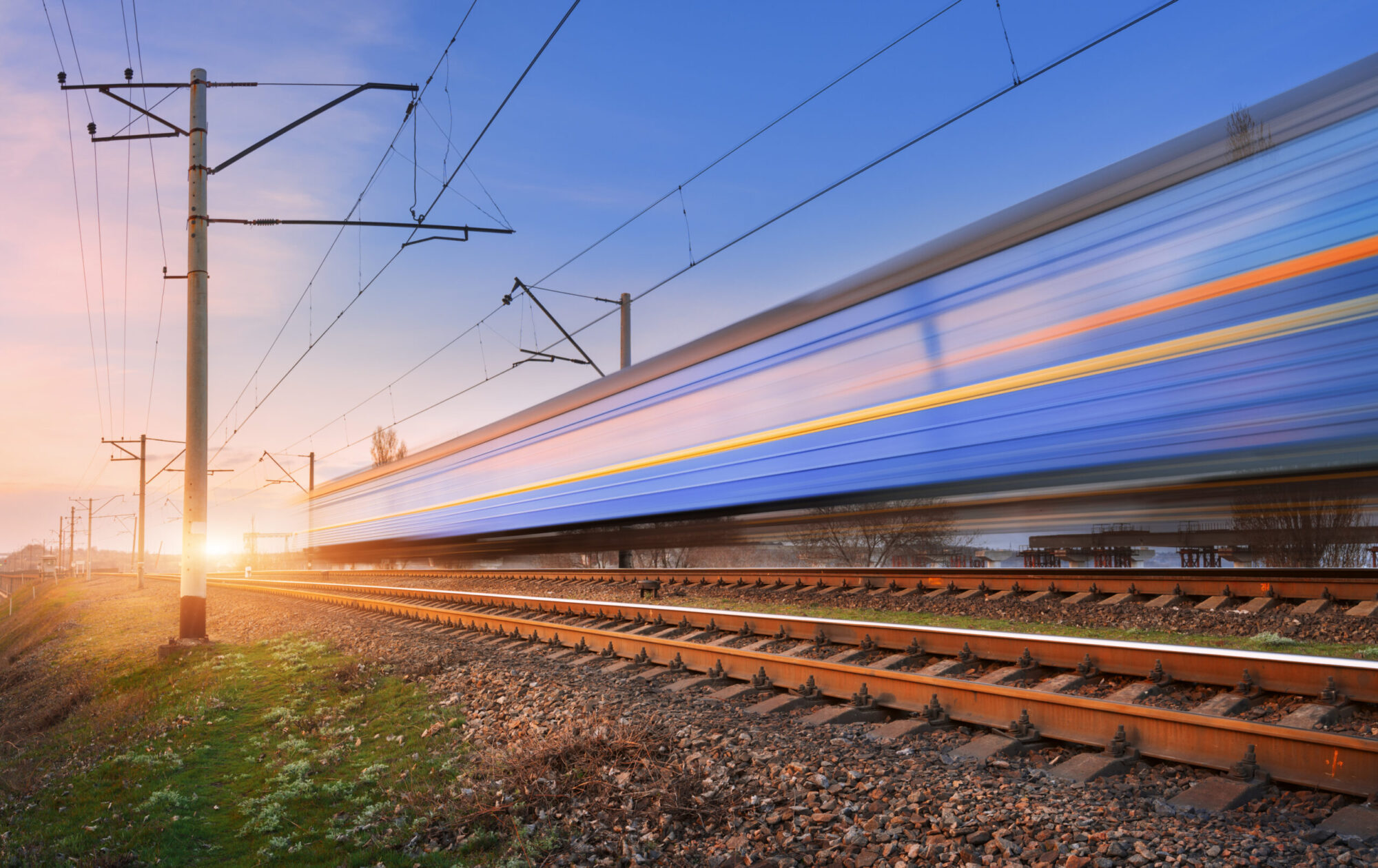 Cette image montre un train en pleine circulation à très grande vitesse. Le train est volontairement flou, ce qui indique qu’il se déplace très rapidement, tandis que les rails, les poteaux et les lignes électriques sont nets : ce contraste sert à renforcer l’idée de mouvement. La scène se déroule sur une voie ferrée électrifiée, visible grâce aux câbles aériens et aux pylônes. La lumière basse, proche du lever ou du coucher du soleil, donne des couleurs chaudes à l’horizon.