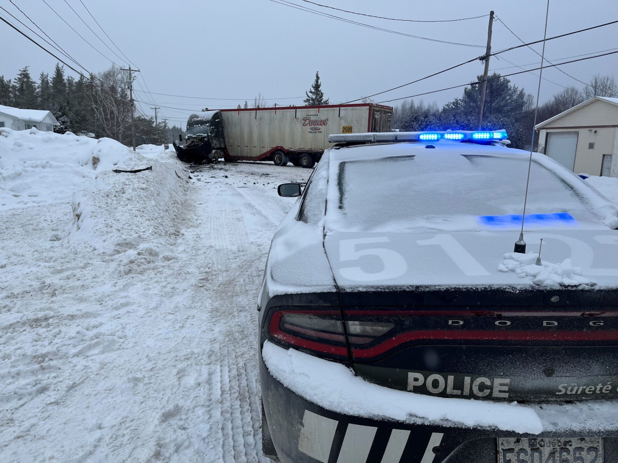Une voiture de police enneigée, gyrophares bleus allumés, est arrêtée au premier plan sur une route couverte de neige. Plus loin, un camion semi-remorque est accidenté en travers de la chaussée, bloquant complètement le passage.