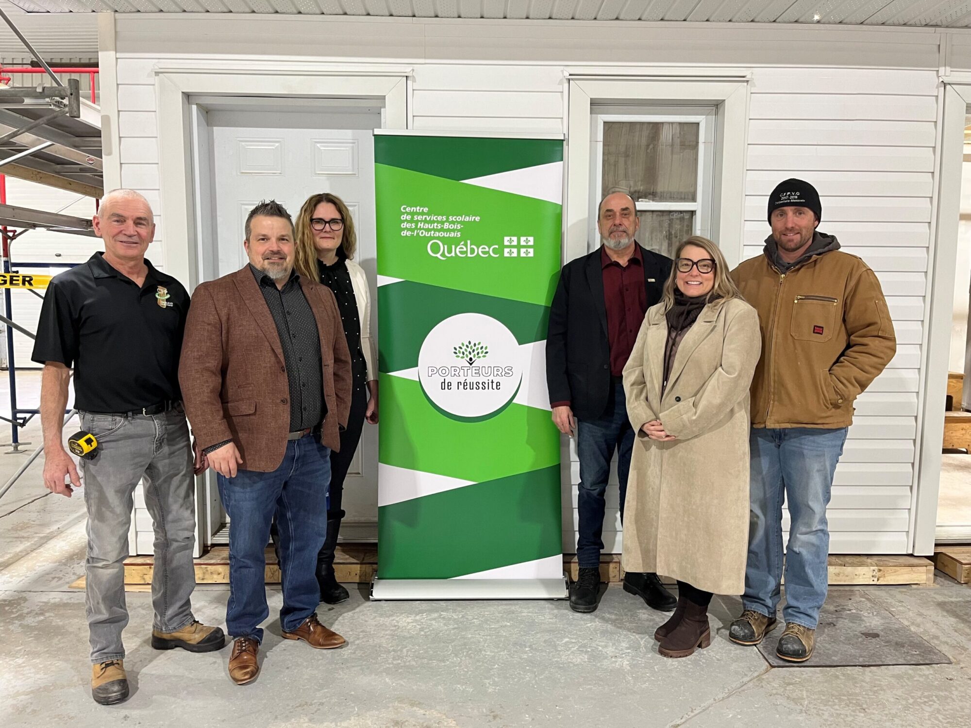 Un groupe de personnes pose à l’intérieur d’un bâtiment qui ressemble à un entrepôt, devant une bannière du Centre de services scolaire des Hauts-Bois-de-l’Outaouais. Derrière eux, se trouve un petit bâtiment en construction.