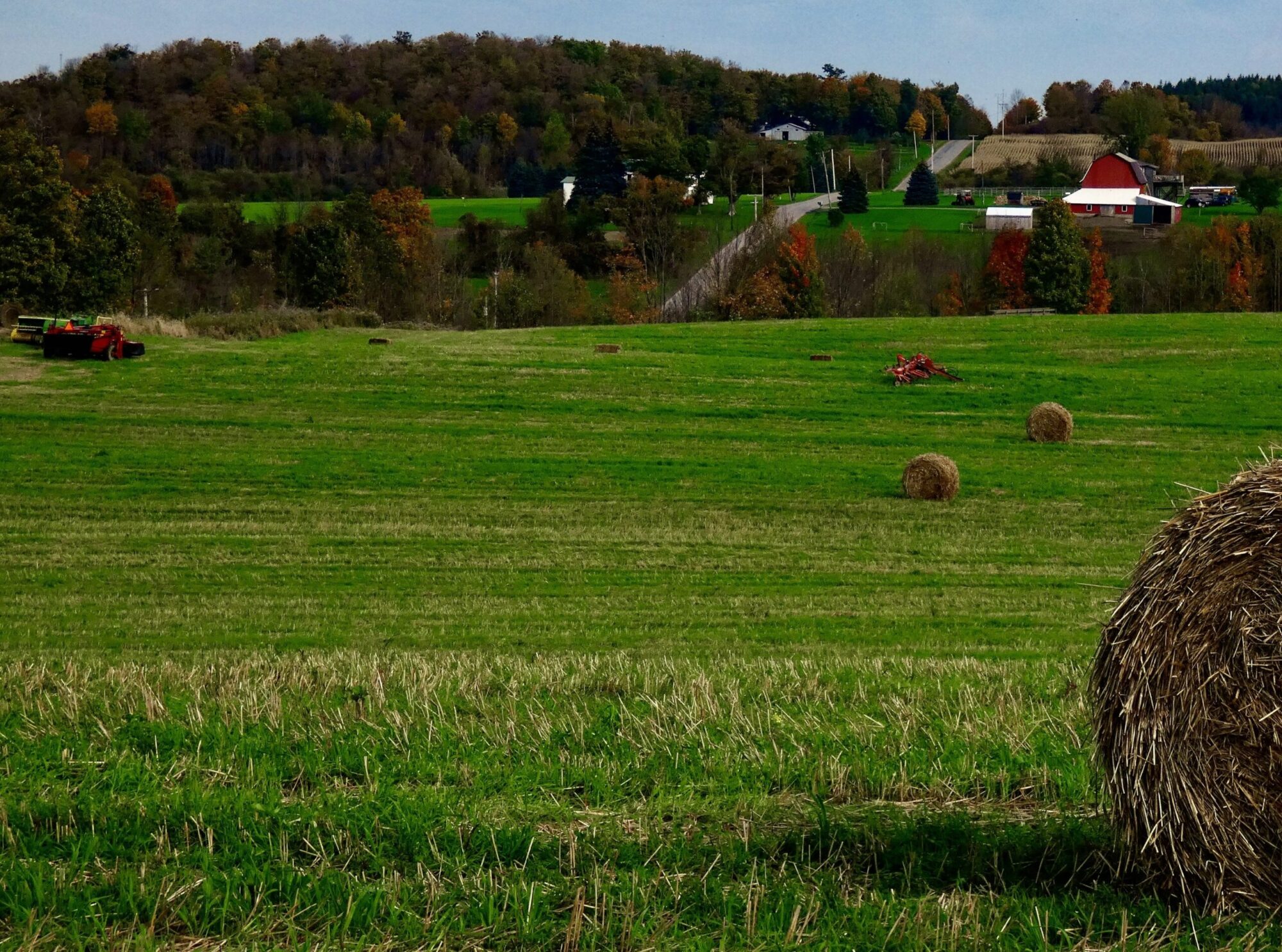 Un champ avec des balles de foin dans un paysage champêtre avec une ferme et une route à l'horizon.