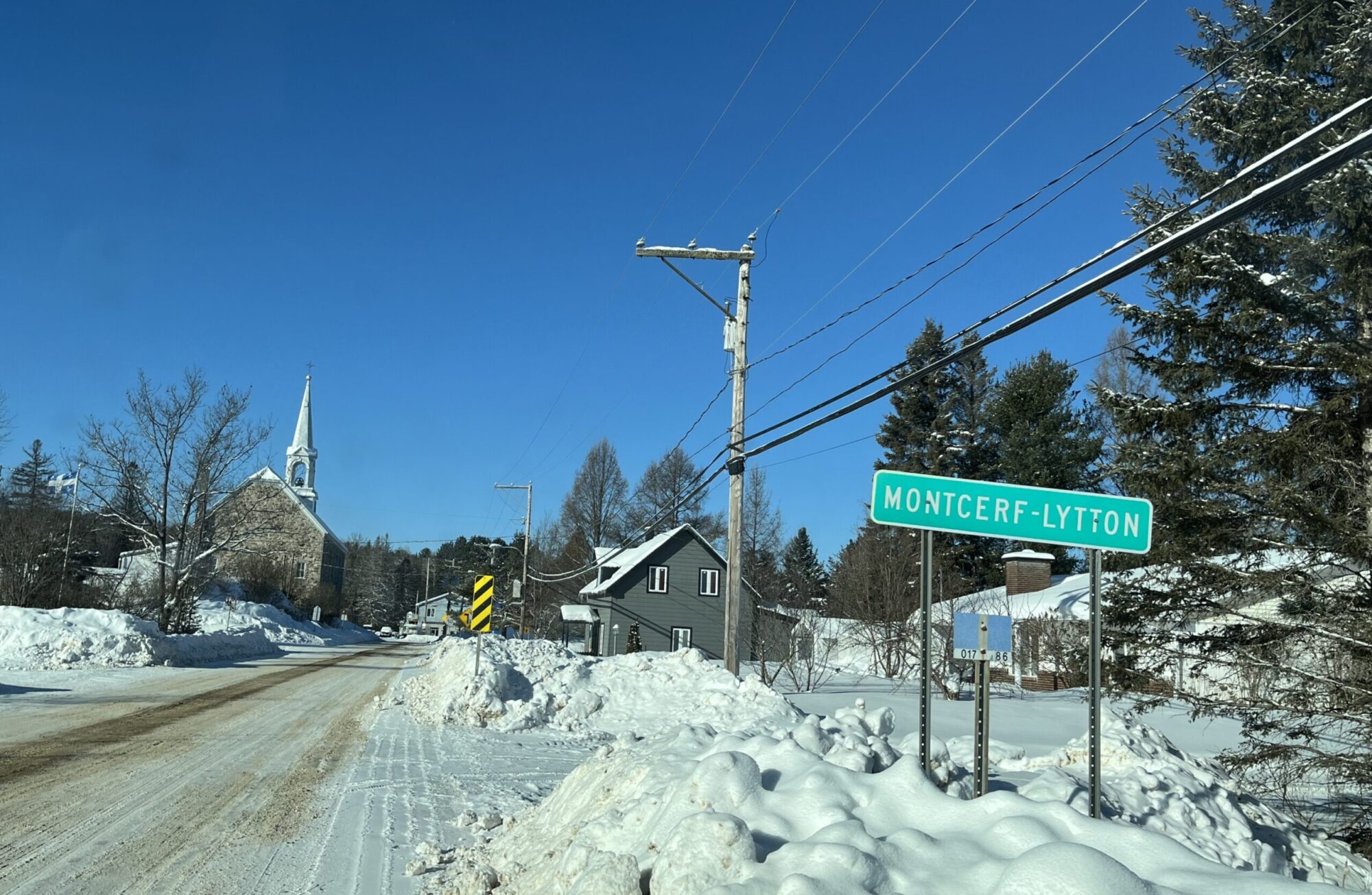 L'entrée d'un village au Québec au cours d'une journée de janvier glaciale avec un grand ciel bleu. Une route enneigée près de laquelle un panneau sur lequel le nom de Montcerf-Lytton est inscrit mène au cœur du village où l'on aperçoit l'Église et des maisons.