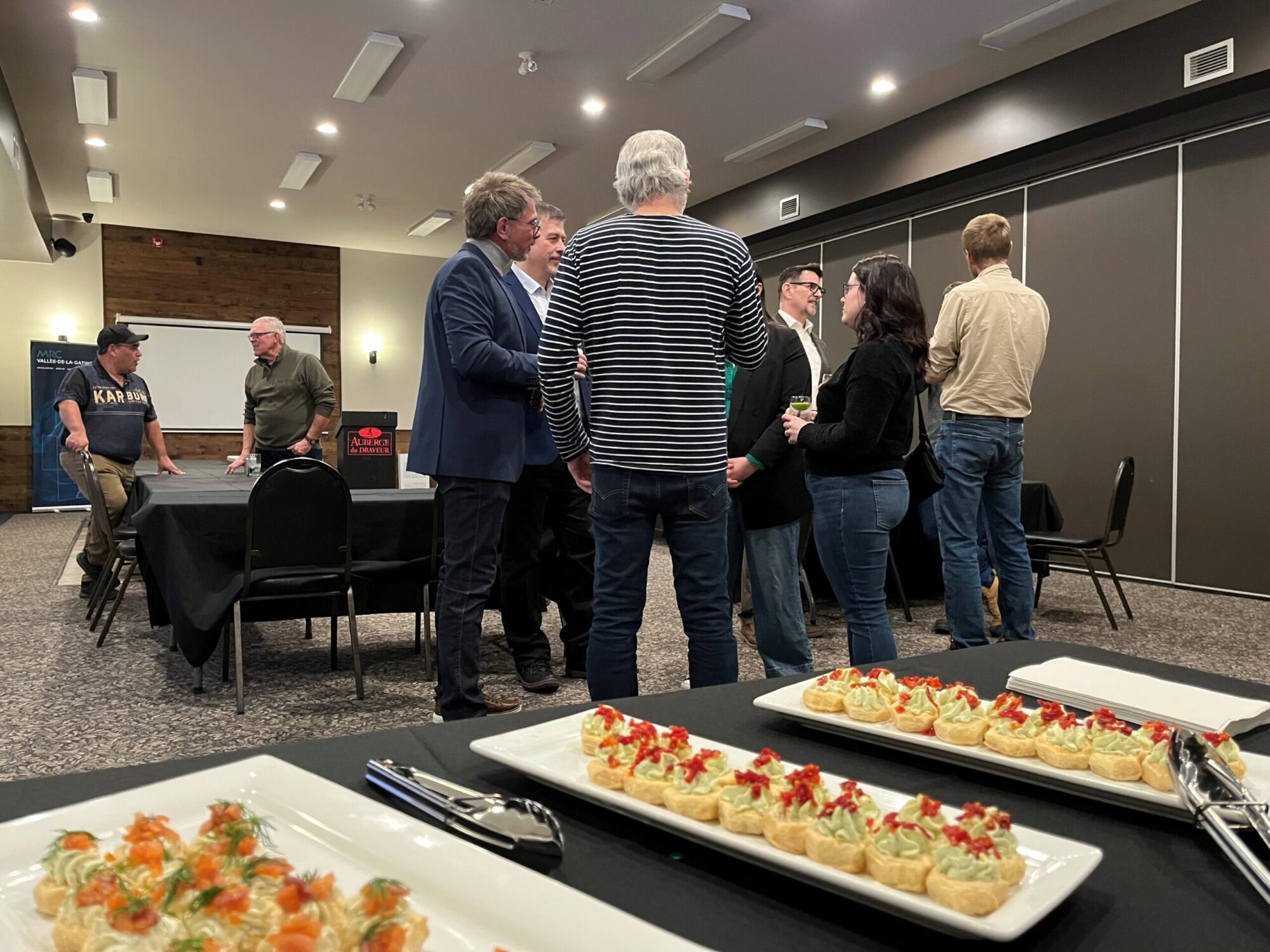 Dans une salle de réception aux murs griss, plusieurs adultes discutent en petits groupes autour de tables recouvertes de nappes noires. Au premier plan, sur une table, des plateaux de bouchées élégantes sont soigneusement disposés à côté de pinces de service.