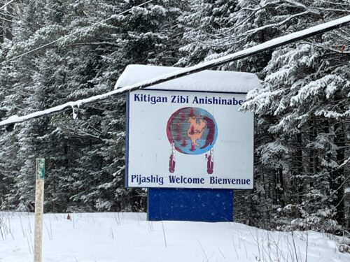 Un panneau bordé de bleu, couvert de neige, se dresse en bordure d’une route forestière enneigée. On y lit « Kitigan Zibi Anishinabeg » au-dessus d’un symbole circulaire représentant une carte et des plumes, ainsi que « Pijashig Welcome Bienvenue », entouré d’arbres chargés de neige et traversé par des fils électriques.