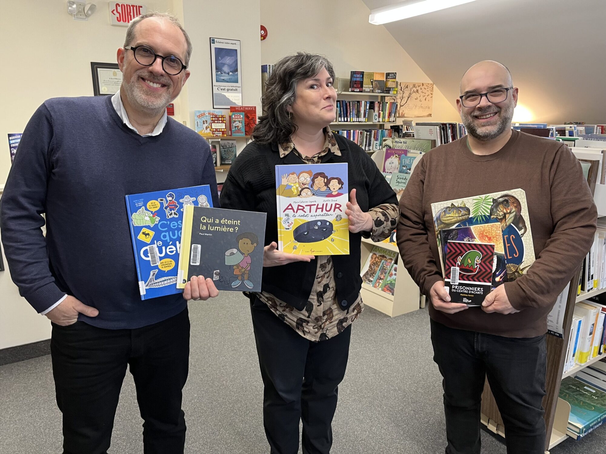 Deux hommes et une femme tiennent des livres dans leurs mains au centre d'un espace de lecture dans une bibliothèque publique.