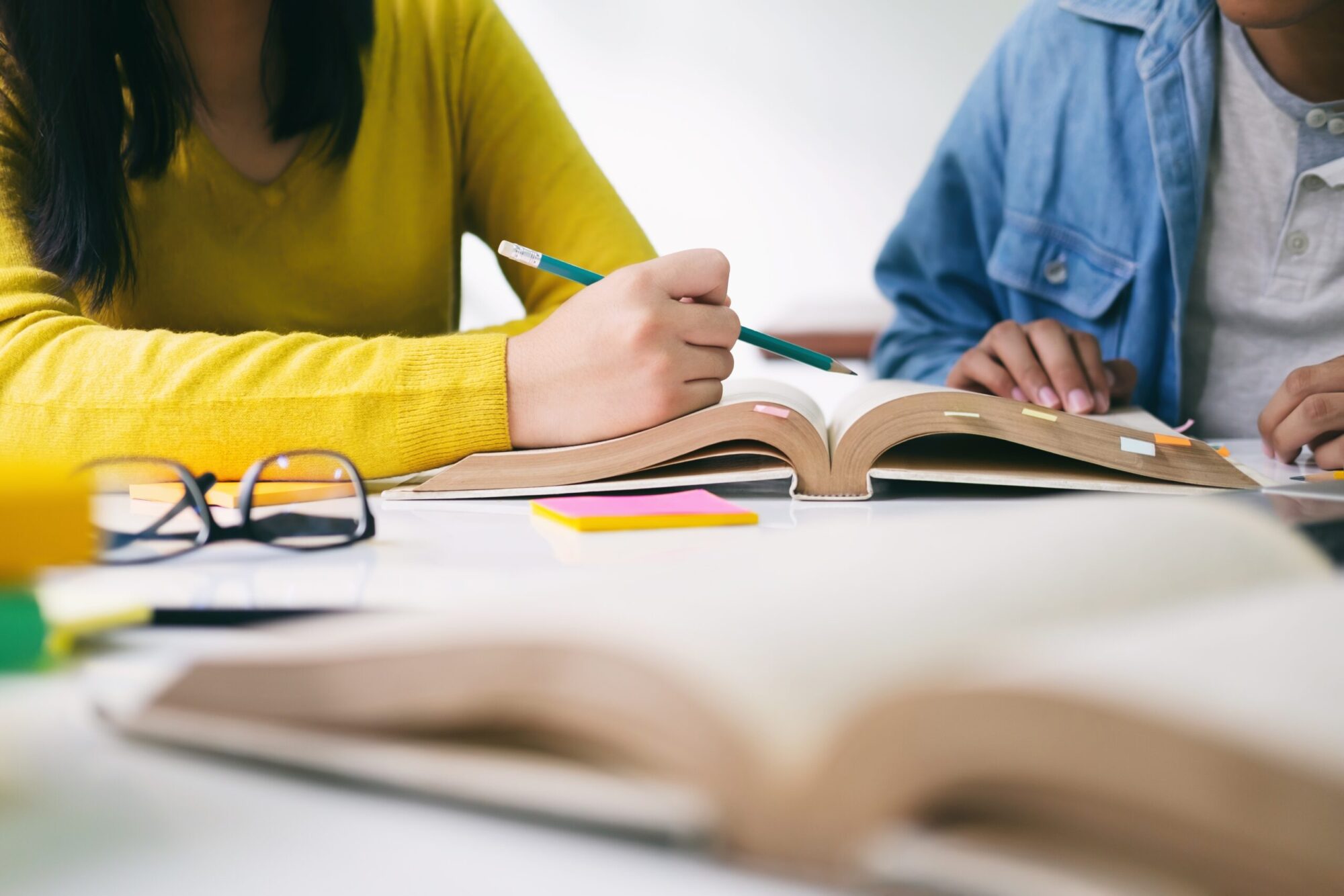 Une personne en chandail jaune pointe du doigt un livre ouvert sur une table avec un crayon à mine, alors qu'une seconde personne en chemise bleue tient le livre. Un autre livre est ouvert à l'avant-plan et des objets comme des post-it et une paire de lunettes sont aussi posés sur la table.