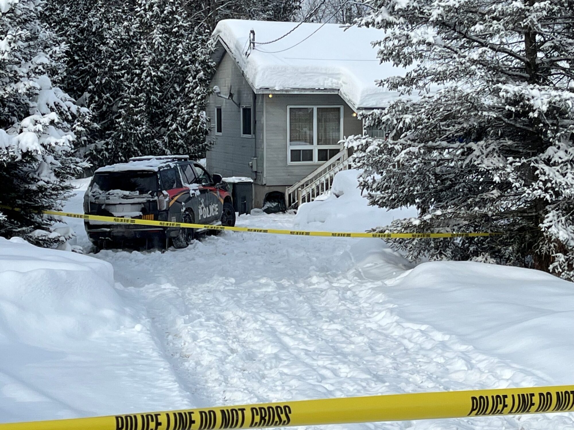 Une petite maison grise est entourée d’arbres chargés de neige, au bout d’une entrée complètement enneigée. Un véhicule de police est stationné devant, et un ruban jaune « Police Line Do Not Cross » bloque l’accès,