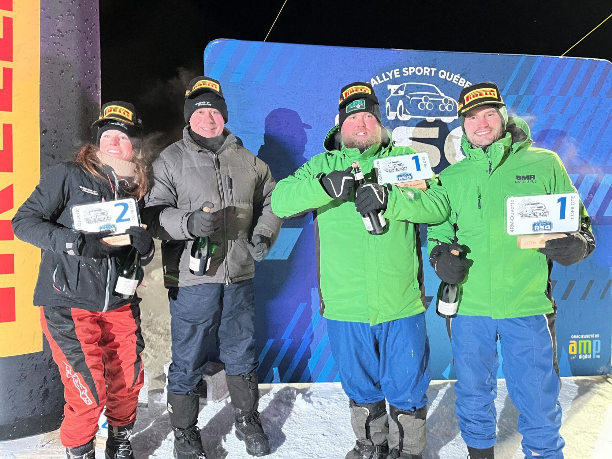 Quatre personnes emmitouflées dans de gros manteaux d’hiver posent sur un podium enneigé, souriantes, tenant des trophées et des bouteilles de champagne, certaines portant des tuques noires et des manteaux verts ou foncés. Derrière elles, un grand panneau bleu de rallye illuminé par la lumière des appareils photo tranche avec la nuit noire.