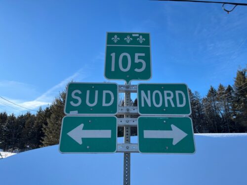 Un panneau routier indique blanc sur vert la route 105, avec trois fleurs de lys au sommet. En dessous, deux panneaux montrent « SUD » avec une flèche vers la gauche et « NORD » avec une flèche vers la droite, devant un paysage enneigé et boisé sous un ciel bleu clair.