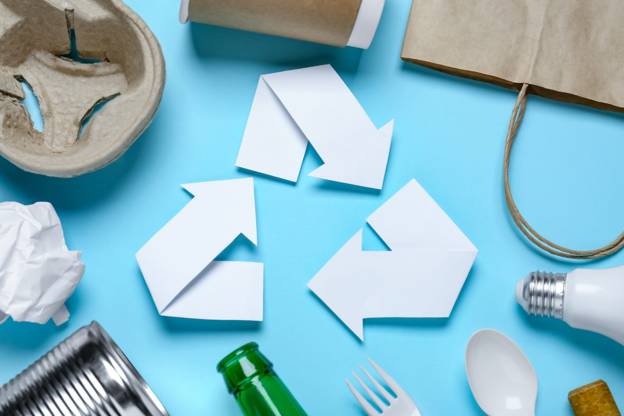 Un symbole de recyclage fait de morceaux de papier blanc posé sur un fon bleu ciel et entouré de divers objets, comme des ustensiles en plastique, du carton et une boîte de conserve.