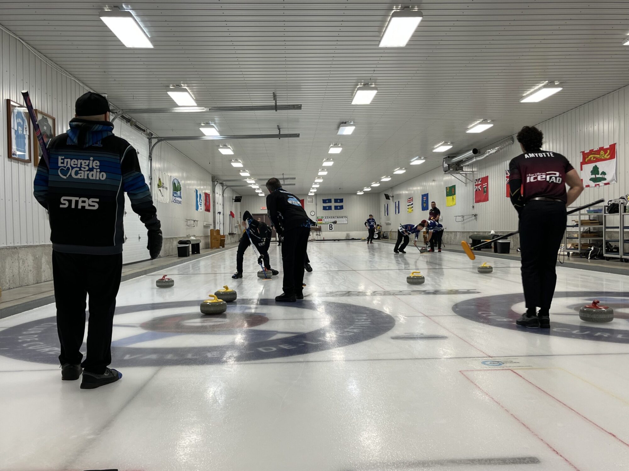 On voit une partie de curling dans un aréna intérieur. Plusieurs joueurs sont sur la glace : l’un lance une pierre tandis que deux coéquipiers balaient devant elle avec leurs balais pour en contrôler la trajectoire et la vitesse. Des pierres jaunes et rouges sont disposées sur la surface, près de la cible circulaire (« maison »). D’autres joueurs attendent ou observent en arrière-plan. Les murs sont décorés de drapeaux et l’éclairage au plafond illumine la patinoire.
