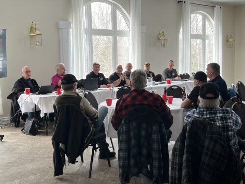 Un groupe d’hommes, principalement âgés, réunis autour de tables dans une salle lumineuse. Ils semblent participer à une réunion ou une discussion, avec des ordinateurs, des papiers et des boissons posés devant eux. L’ambiance est calme et attentive, comme lors d’une assemblée ou d’un comité.