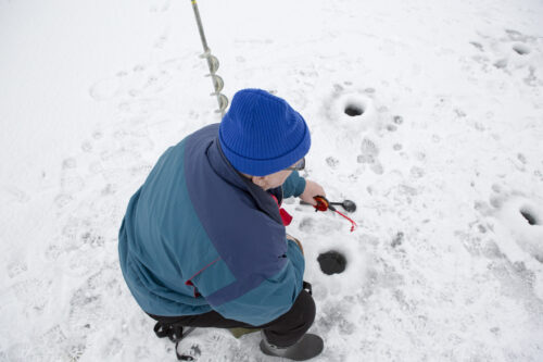 Un vieil homme en manteau, bottes et tuque d'hiver pêche sur un lac percé de trous durant la saison froide.