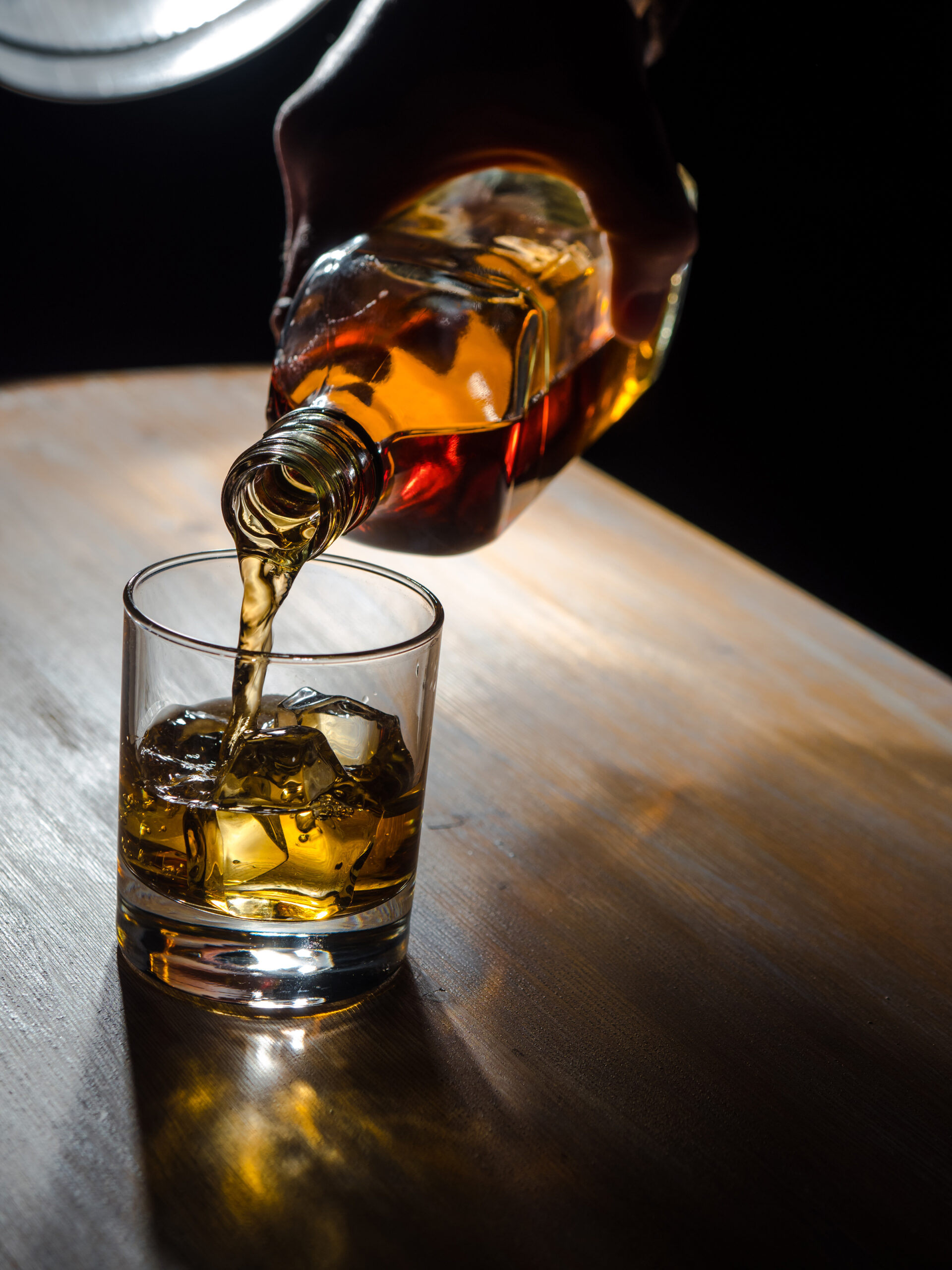 Une main verse de l'alcool d’une bouteille de verre dans un verre tumbler contenant des glaçons. La scène est éclairée de façon dramatique sur une table en bois sombre, créant des reflets dorés et des ombres marquées.