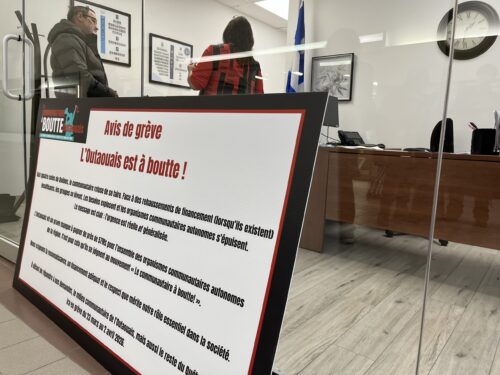 Une pancarte qui menace de grève est posée contre la vitre d'un bureau de circonscription dans lequel des personnes parlent.