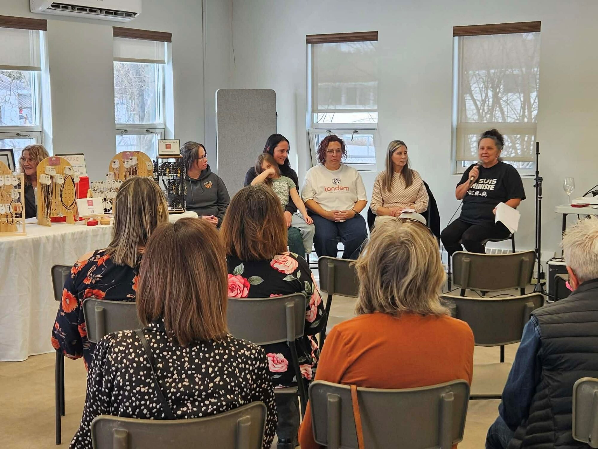 Dans une salle lumineuse, un groupe de femmes est assis face à un public. L’une d’elles, à droite, parle au micro. Les autres l’écoutent attentivement. Au premier plan, plusieurs personnes vues de dos sont assises sur des chaises et assistent à la présentation. À gauche, une table d’exposition présente des bijoux artisanaux.