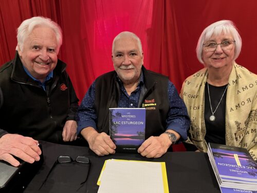 Deux hommes et une femme âgés devant un rideau rouge sont assis derrière une table recouverte d'une nape noire sur laquelle des livres sont mis en vente.