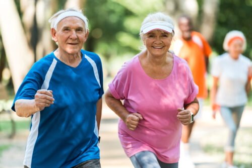 On voit un couple de personnes âgées en train de faire du jogging dans un parc. Ils portent des vêtements de sport et des bandeaux, et ils sourient, donnant une impression d’énergie et de bonne humeur. En arrière-plan, d’autres personnes semblent également faire de l’exercice, dans un environnement verdoyant et ensoleillé.