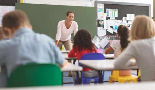 Une professeure s'adresse à sa classe d'enfants de niveau primaire qui l'écoutent en étant assis sur leurs chaises.