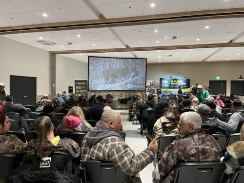 Un public majoritairement vêtu en habits de chasse attend dans une grande salle le début d'un film dédié à la chasse. On voit un cerf de Virginie sur un écran.