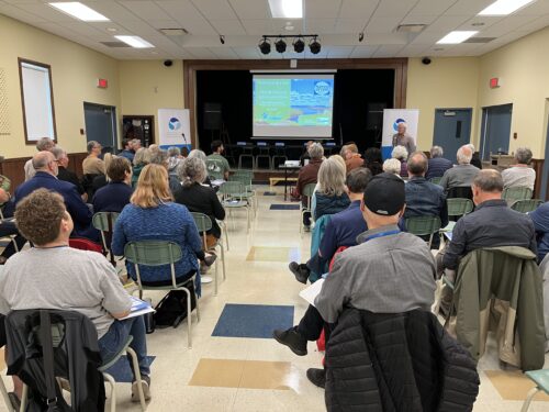 Un conférencier s'adresse à des personnes assises sur des chaises dans une salle communautaire., avec une présentation sur le thème de l'eau sur un écran.