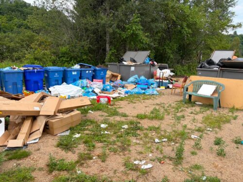 Site de dépôt de matières résiduelles en plein air, avec plusieurs bacs de recyclage bleus, des conteneurs gris et un amas de cartons, sacs de plastique et déchets éparpillés au sol. Des chaises en plastique et du mobilier abandonné se trouvent à droite, devant une zone boisée.