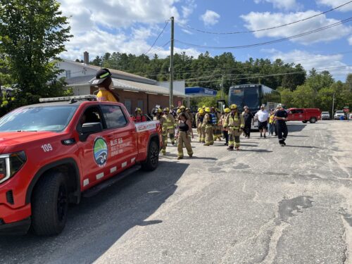 Des pompiers escortés par un de leurs camions et suivis par un autobus marchent en habit de combat dans les rues de Gracefield le 15 août 2026.